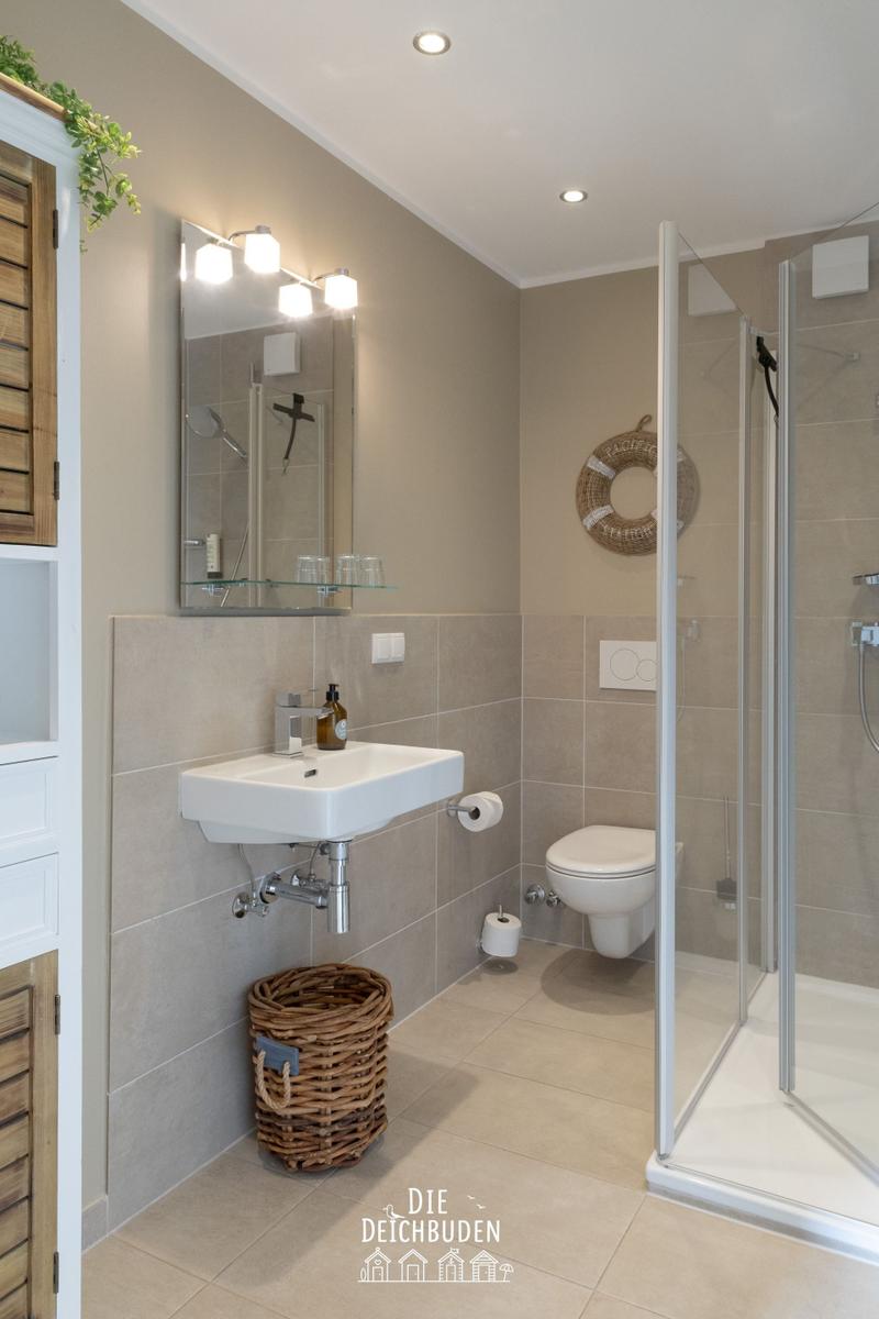 Badezimmer mit Dusche, Waschbecken und Toilette. Holzregal und Korb sind sichtbar.