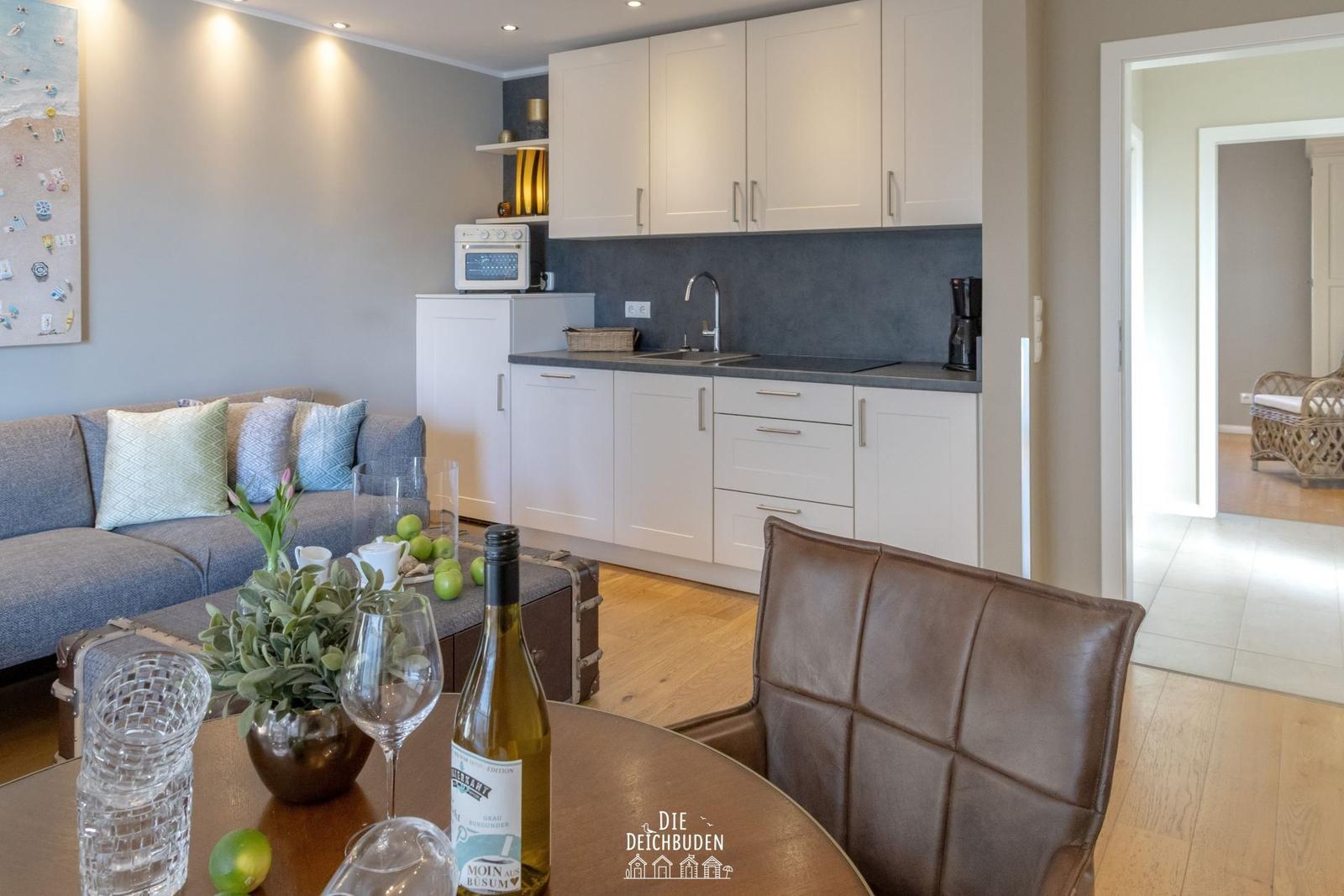 Küche mit weißen Schränken und grauer Arbeitsplatte. Wohnbereich mit Sofa und Esstisch.