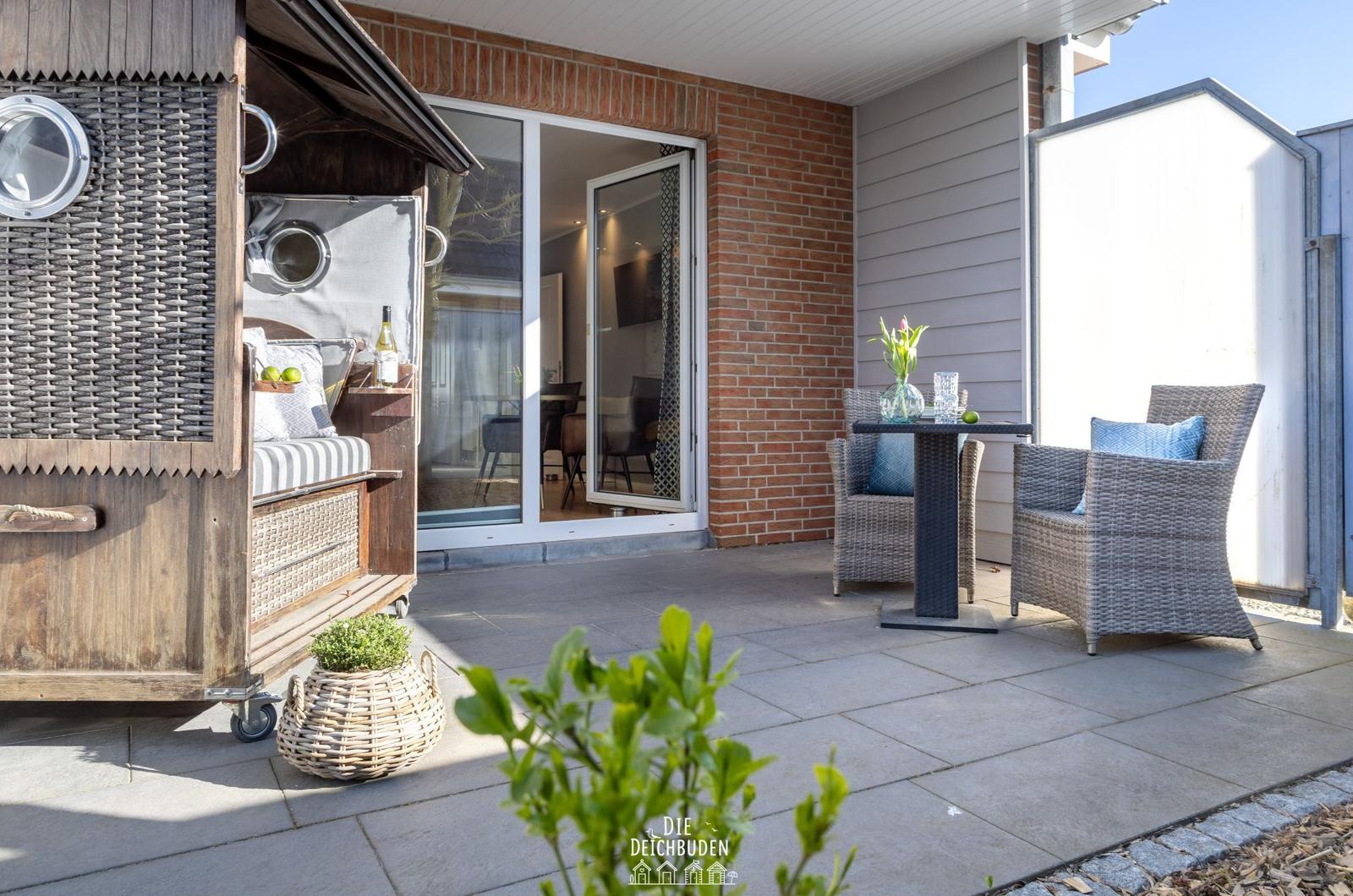 Terrasse mit Sitzgruppe, Glastür und Holzhaus. Pflanzen und Tisch sind sichtbar.