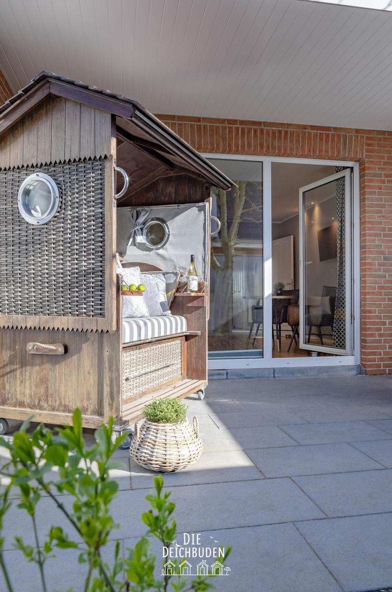 Holzkonstruktion mit Rattan, Sitzbank und Flasche im Freien. Hintergrund: Wohnbereich mit Glasfront.