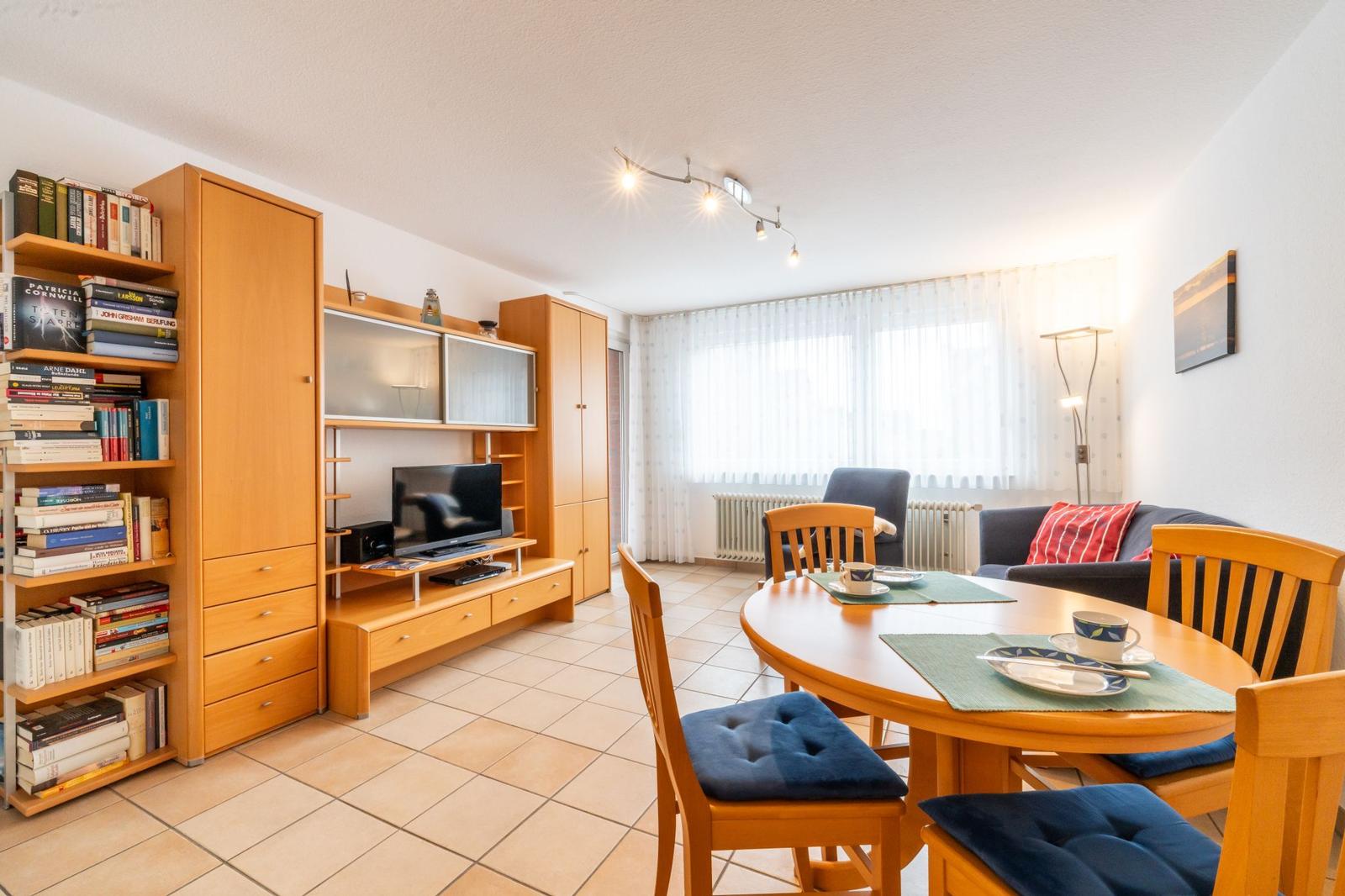 Wohn- und Essbereich mit Holzmöbeln, TV und Bücherregal. Fenster mit weißen Vorhängen.