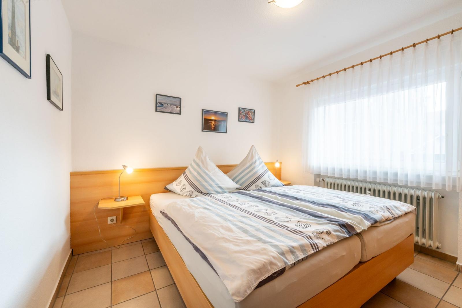 Schlafzimmer mit Doppelbett, Holzgestell, Fenster mit Vorhängen und Nachttisch mit Lampe.