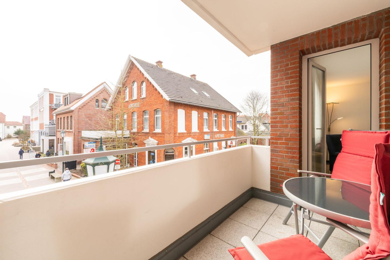 Balkon mit roten Sesseln und Tisch. Blick auf rote Backsteinhäuser mit Apotheke.