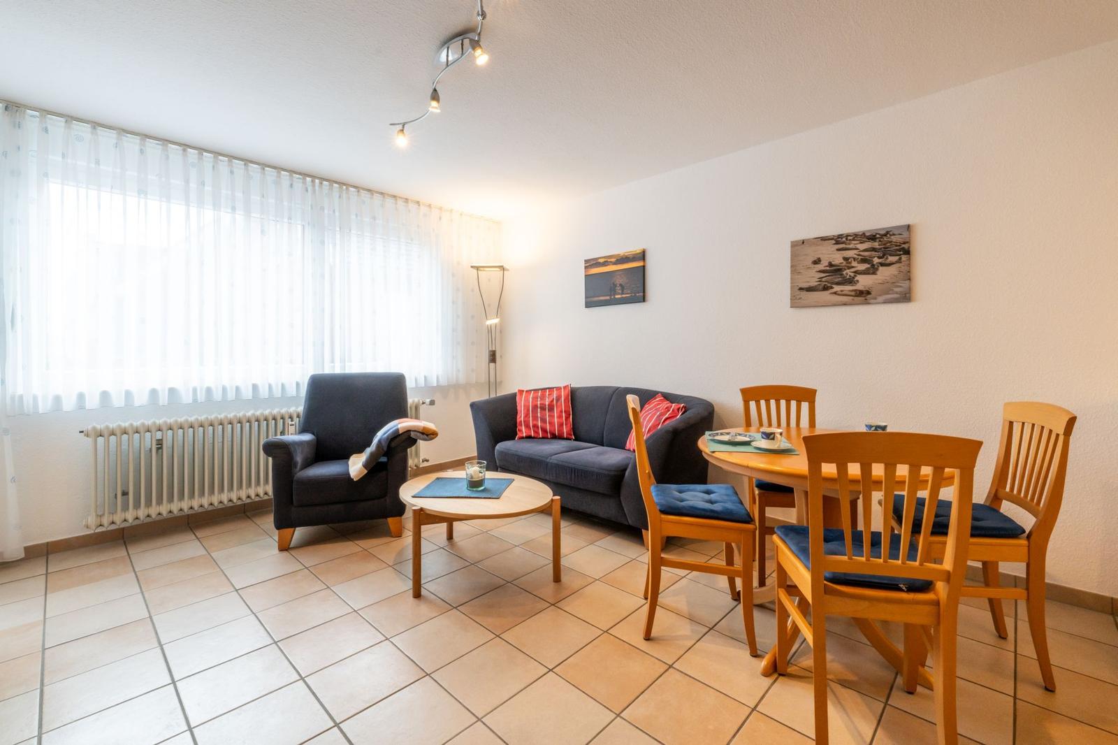 Wohn- und Essbereich mit Sofa, Sessel, Tisch und Stühlen. Fenster mit Vorhängen und Wandbilder.