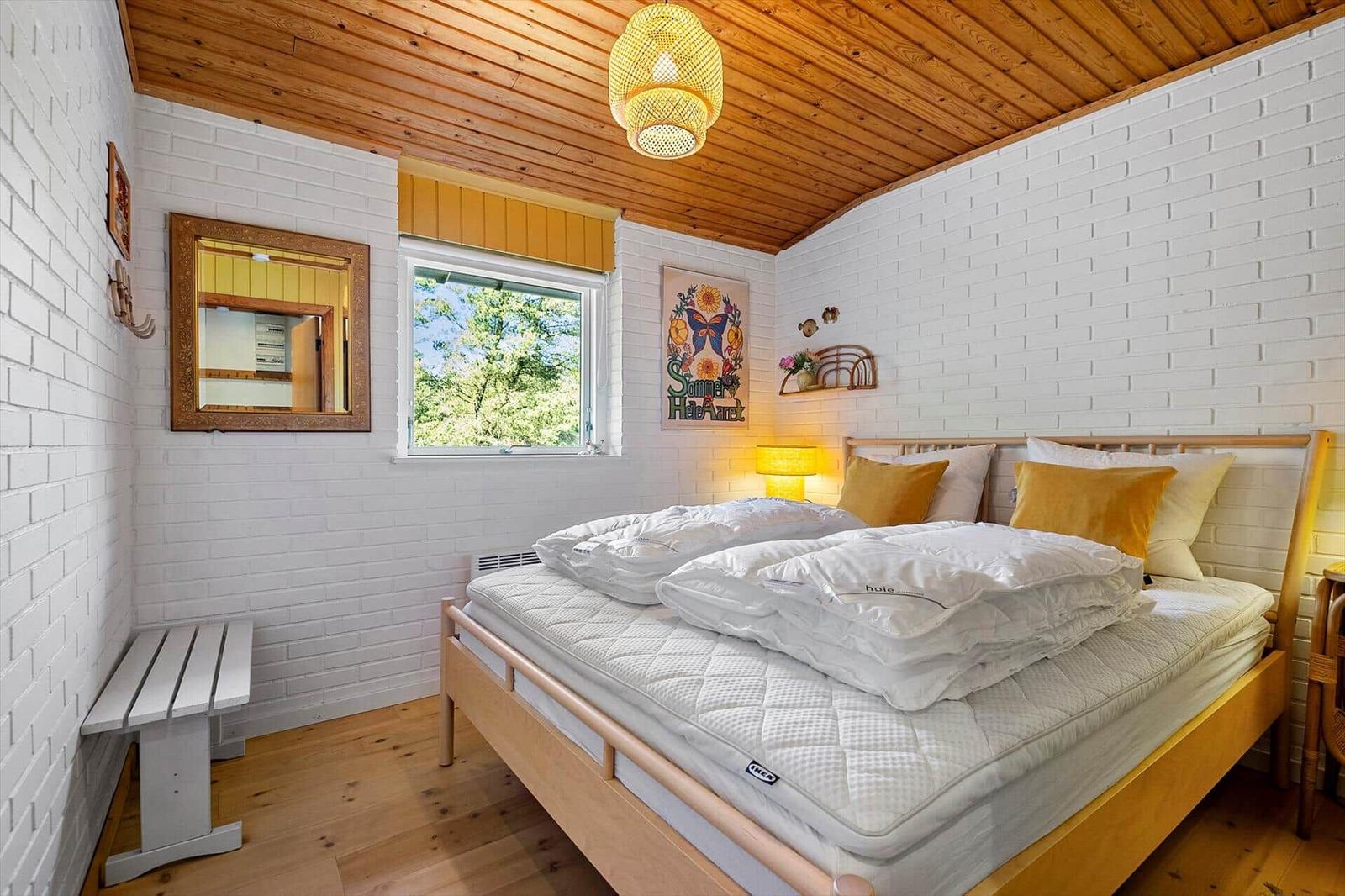 Schlafzimmer mit weißem Bett, Holzdecke und Fenster mit Blick auf Bäume.