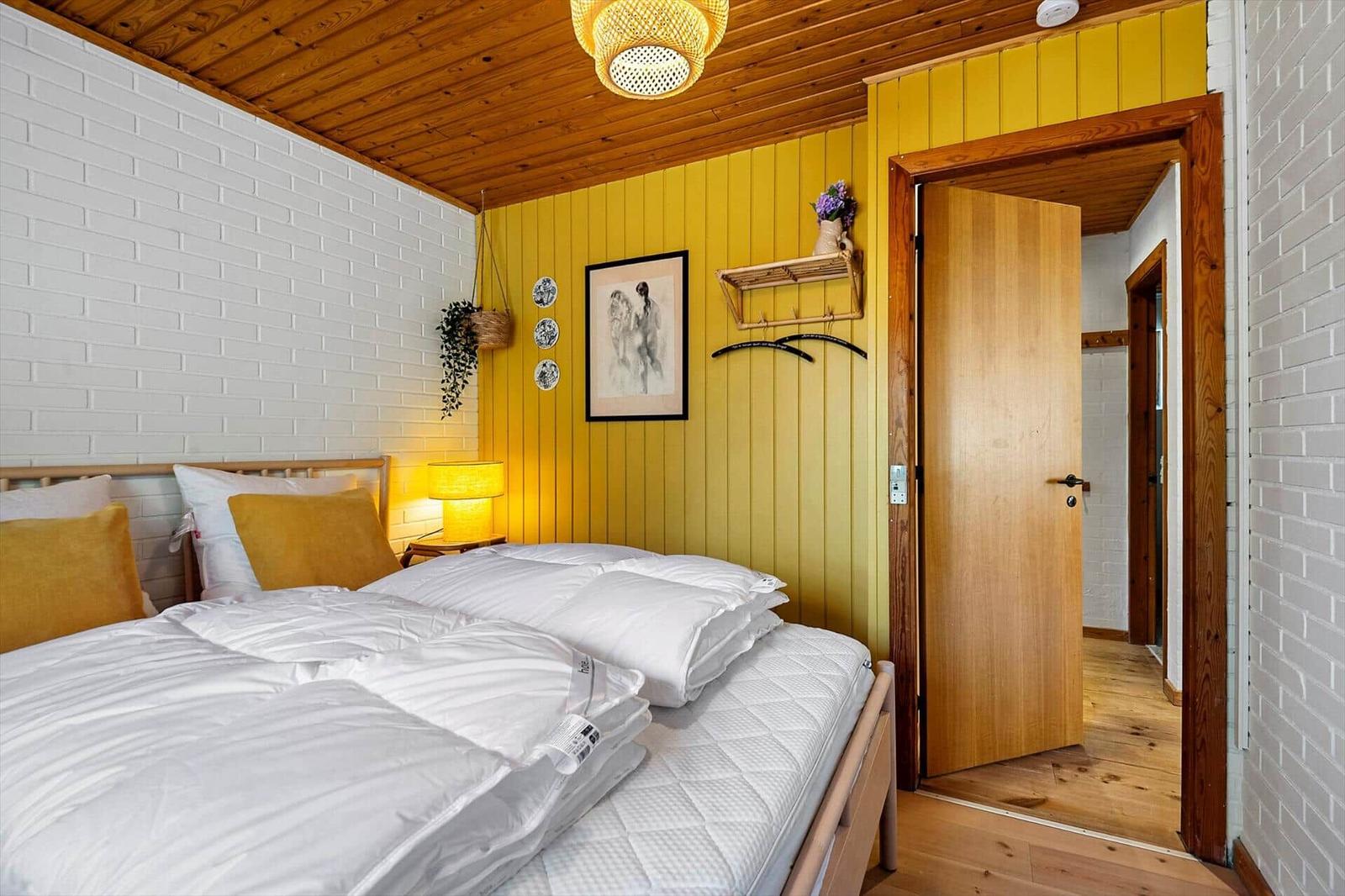 Schlafzimmer mit Bett, gelber Wand und Holzdecke. Tür führt in einen Flur.
