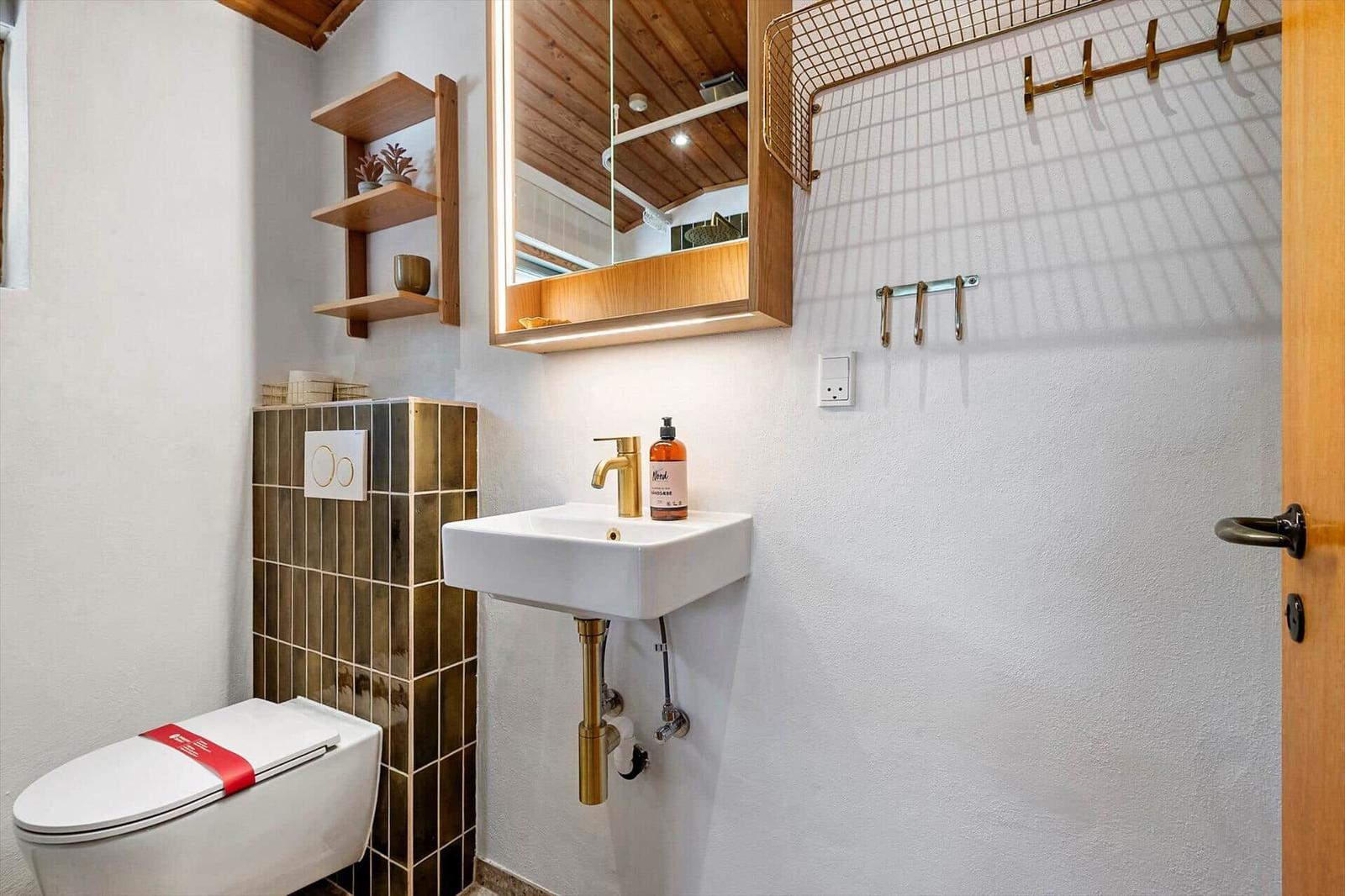 Badezimmer mit Toilette, Waschbecken und Spiegelschrank. Holzregale und goldfarbene Armaturen.