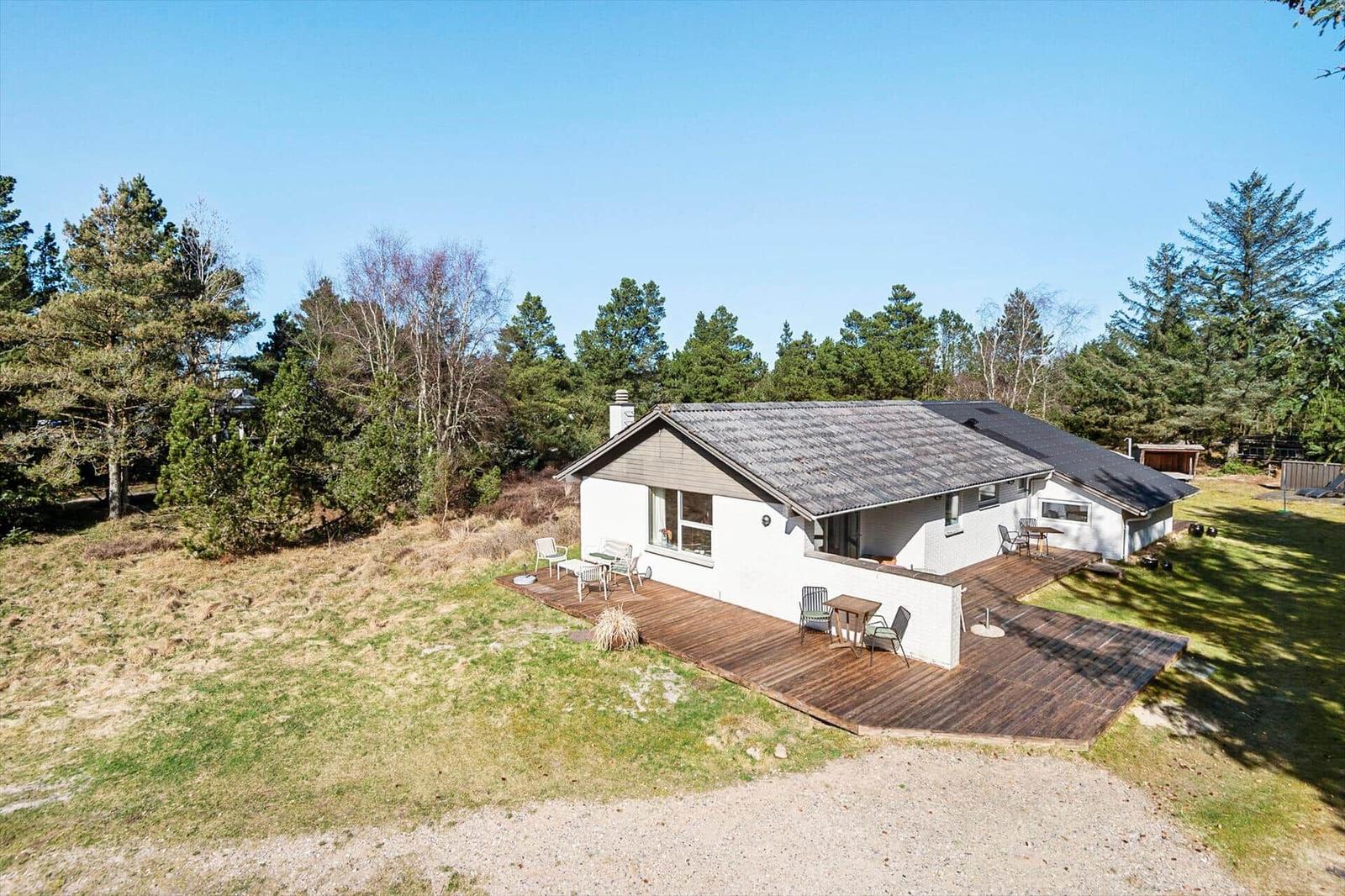 Ein Haus mit Holzterrasse und Garten, umgeben von Bäumen unter blauem Himmel.
