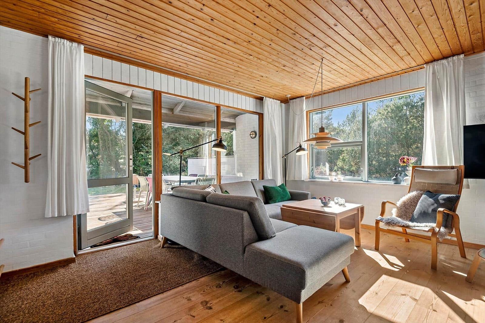 Wohnzimmer mit Sofa, Sessel und Holzboden. Große Fenster und Glastüren bieten Blick auf Garten.