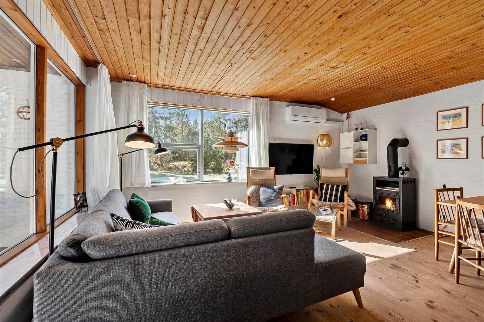Wohnzimmer mit Holzdecke, Sofa, Kamin und großem Fenster. Essbereich mit Holztisch und Stühlen.