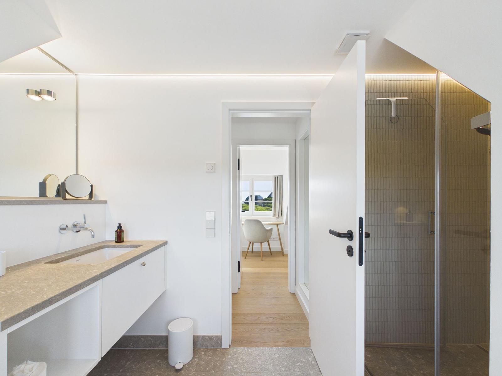Badezimmer mit Dusche und Waschbecken. Blick in Wohnbereich mit Sitzgelegenheit.