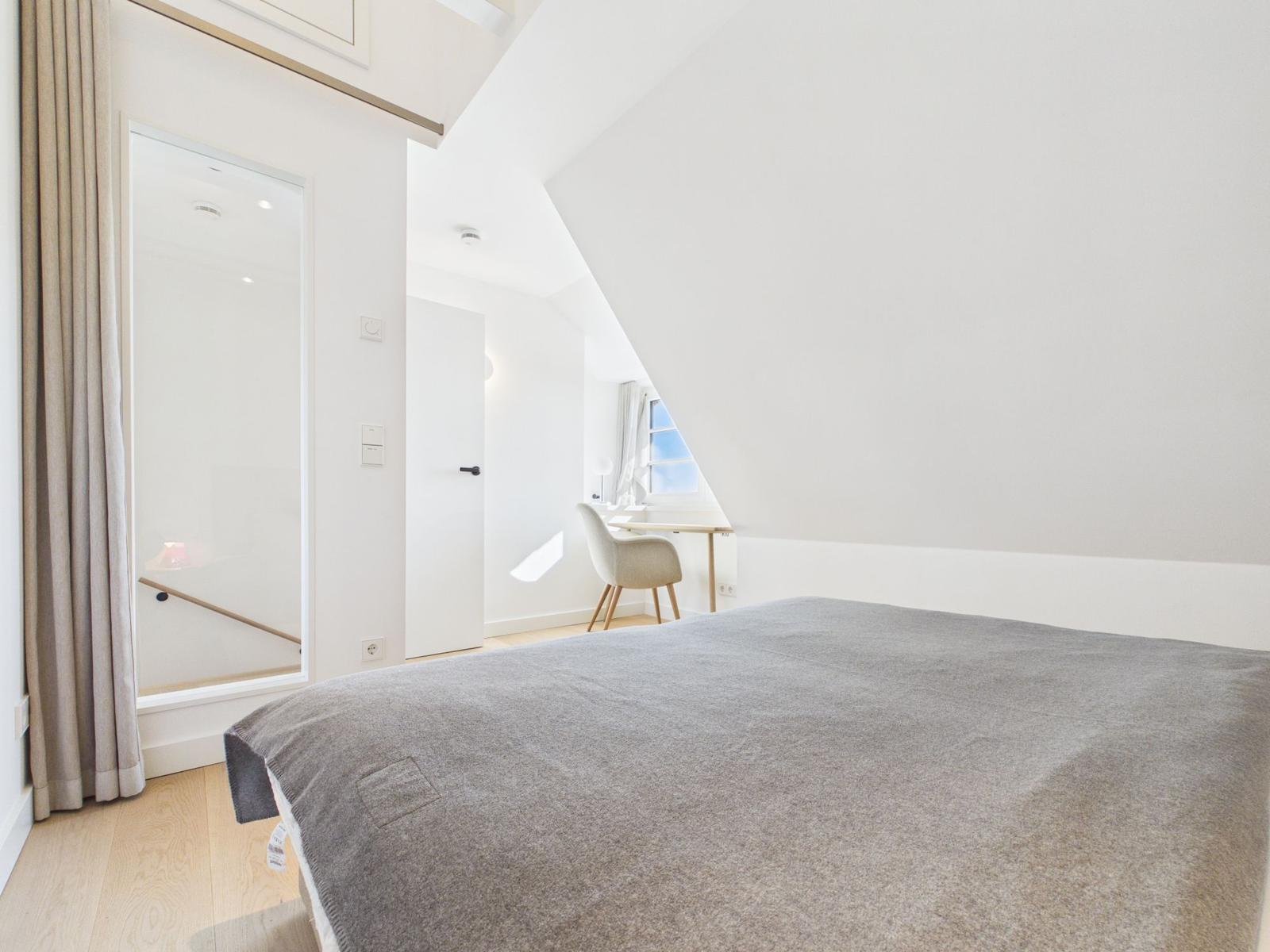 Schlafzimmer mit Bett, Schreibtisch und Fenster. Dachschrägen und helle Wände.