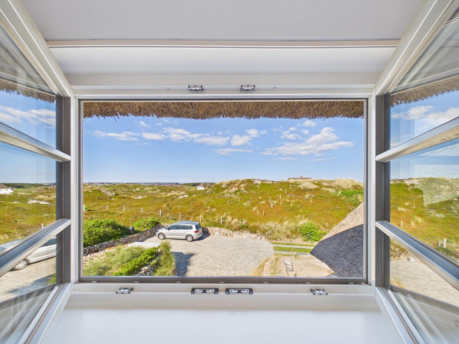 Durch das Fenster blickt man auf eine grüne Dünenlandschaft mit einem geparkten Auto.