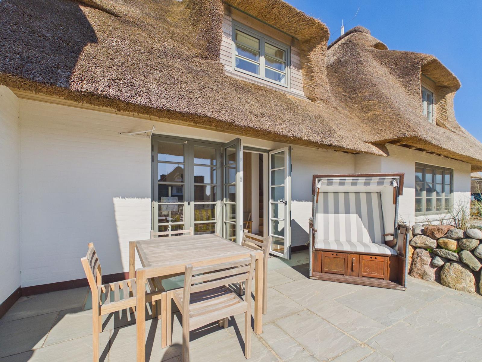 Terrasse mit Tisch, Stühlen und Strandkorb vor einem Haus mit Strohdach.