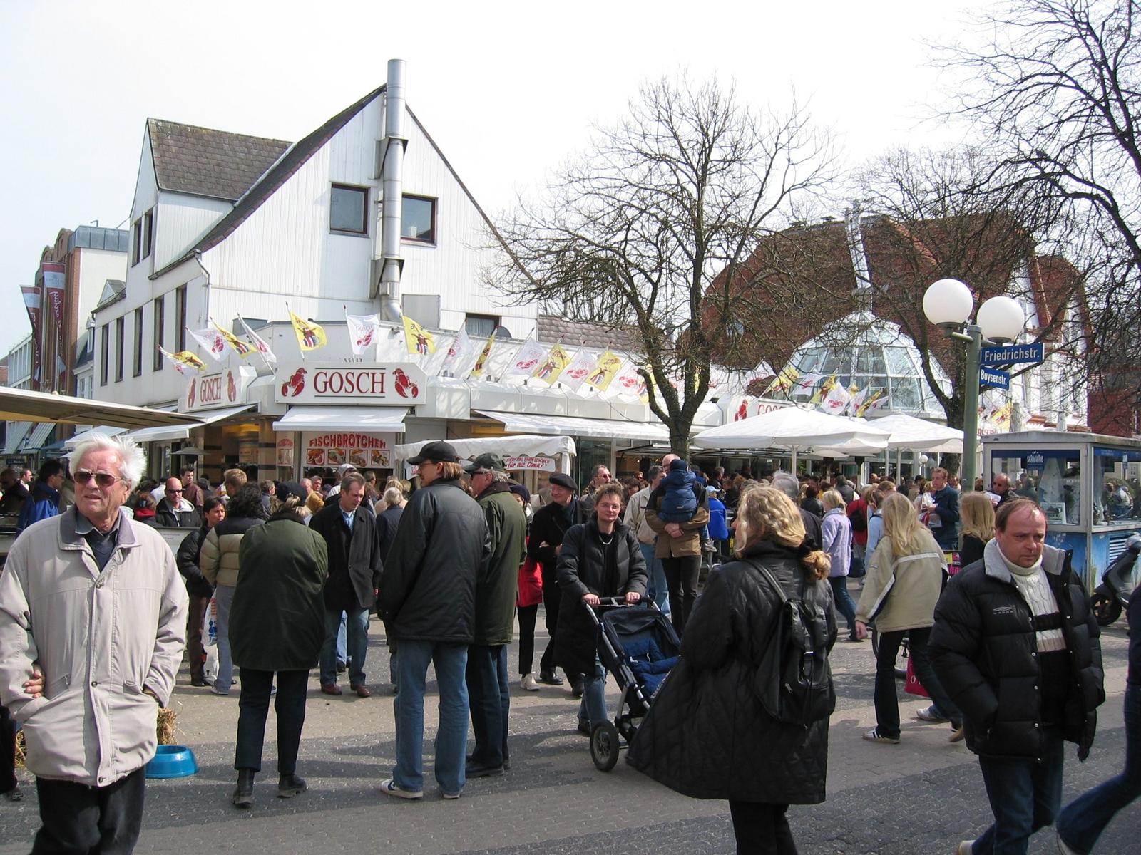 Many people move around a busy street with market stalls and a white building.