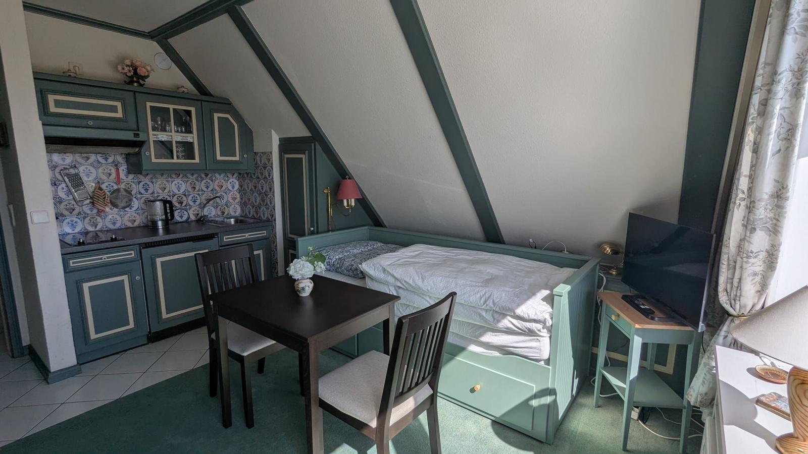 Bedroom with bed, table, chairs, and small kitchen under sloped ceiling.