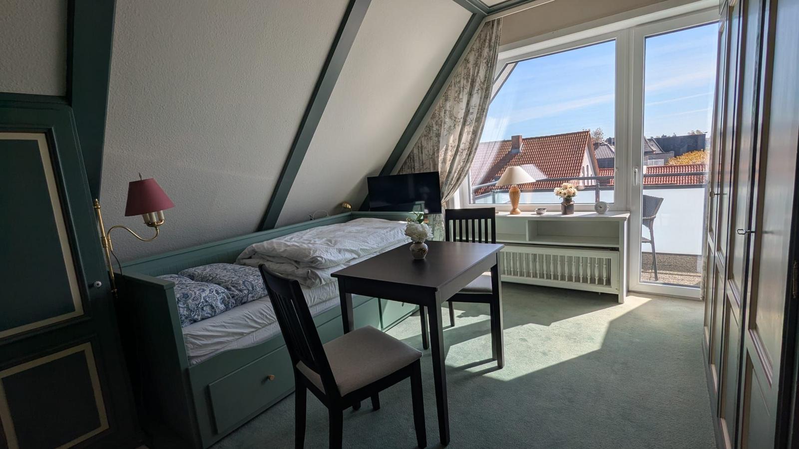 Room with bed, table and chairs, view onto balcony with sight on rooftops.