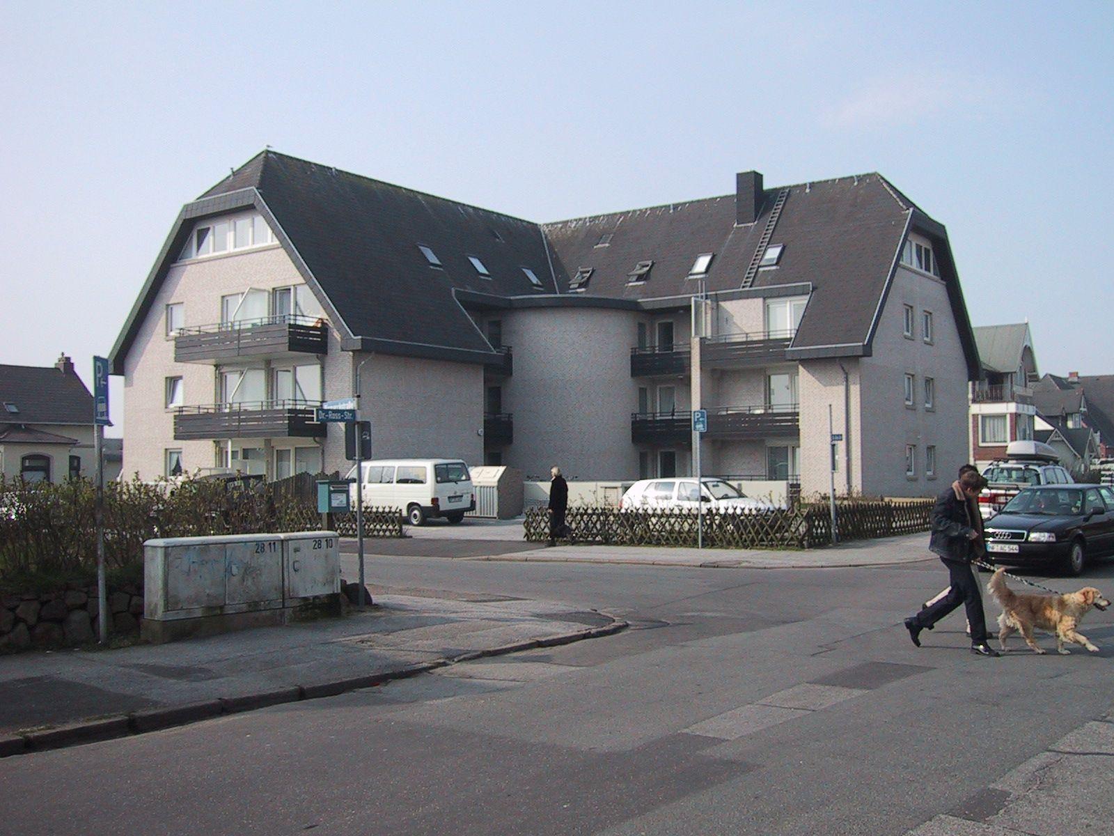 Modern multi-family house with balconies and paved courtyard. A man walks a dog across the street.