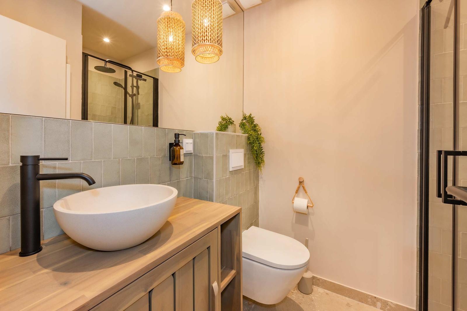 Badezimmer mit Waschbecken, Toilette und Dusche. Holzunterschrank und zwei Hängelampen.