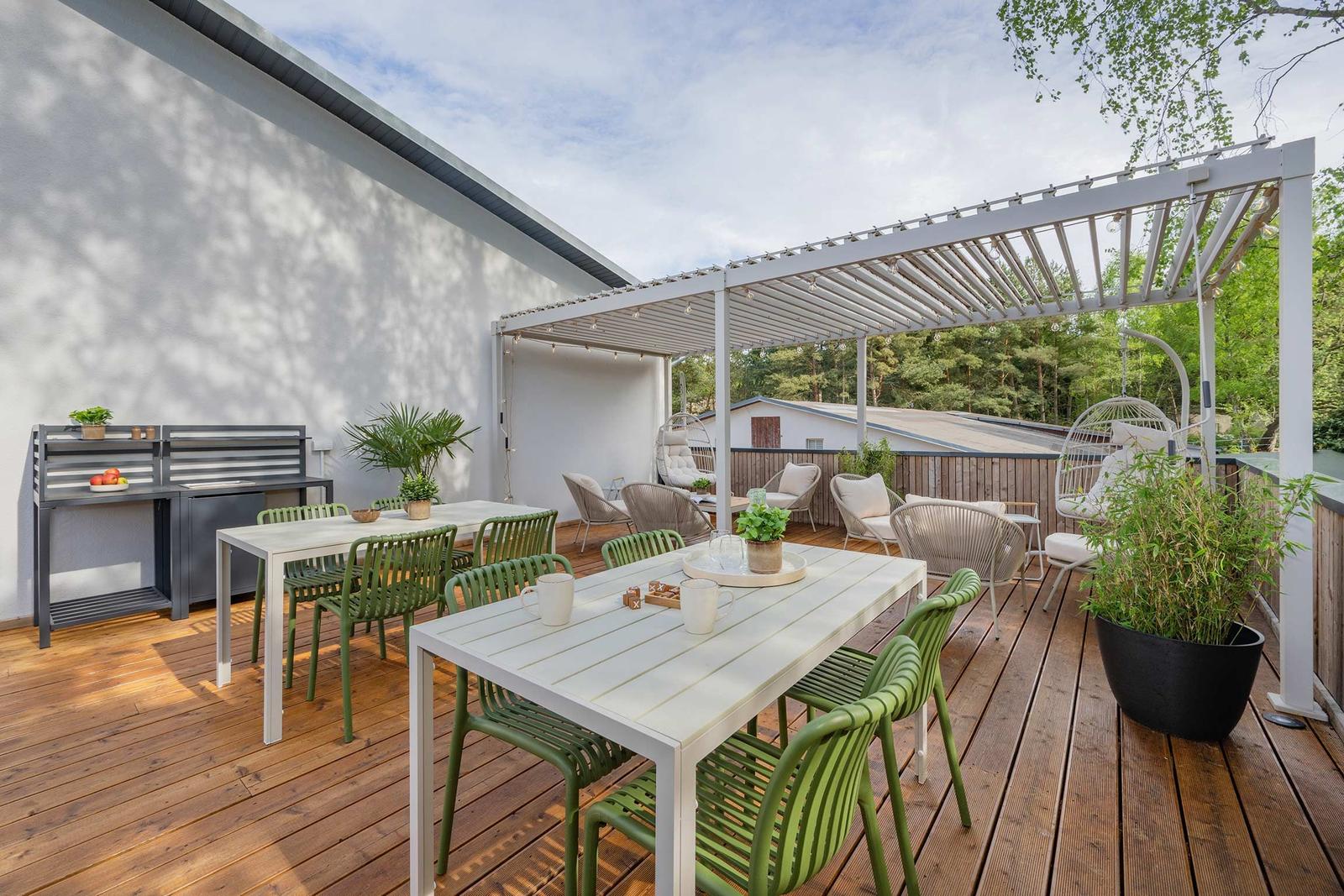 Terrasse mit Tischen, Stühlen und Pergola. Holzboden und Pflanzen sichtbar.