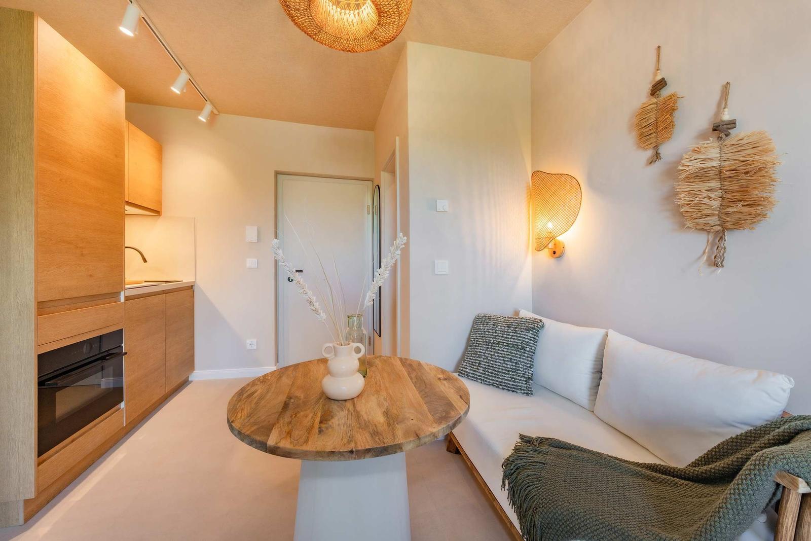 Wohnbereich mit Sofa, runden Tisch und Küchenzeile mit Holzmöbeln.