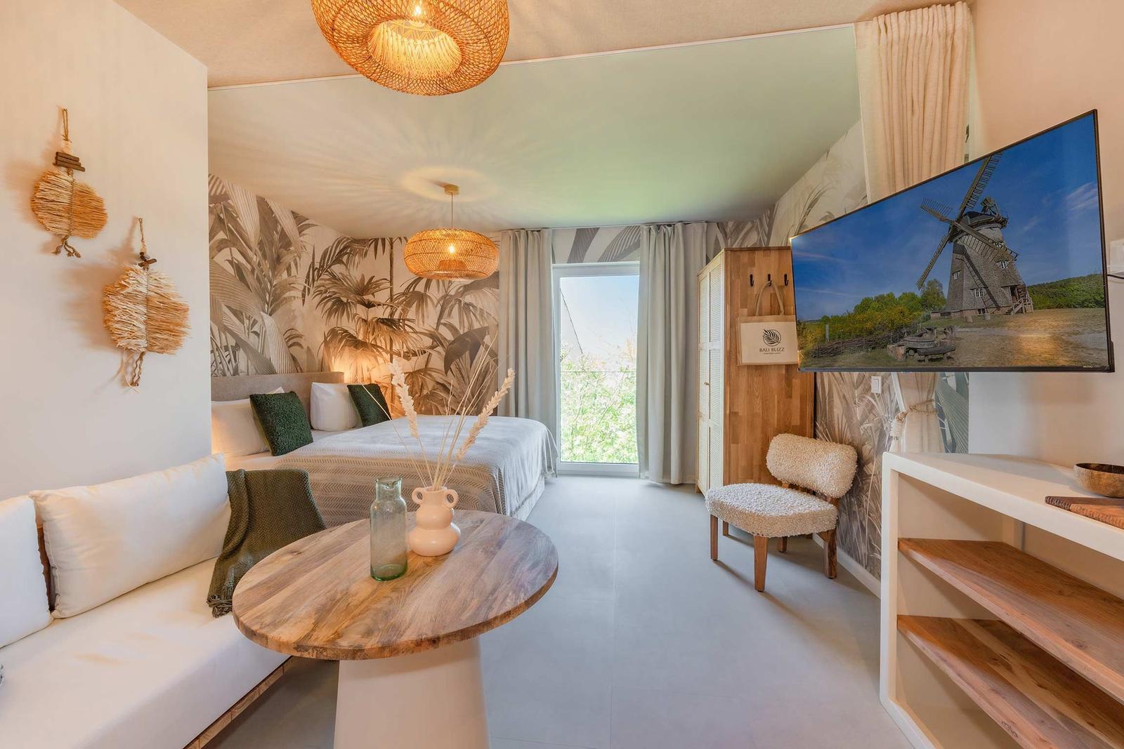 Schlafzimmer mit Bett, Sitzbank, Tisch und Fernseher. Wand mit Palmenmotiv.