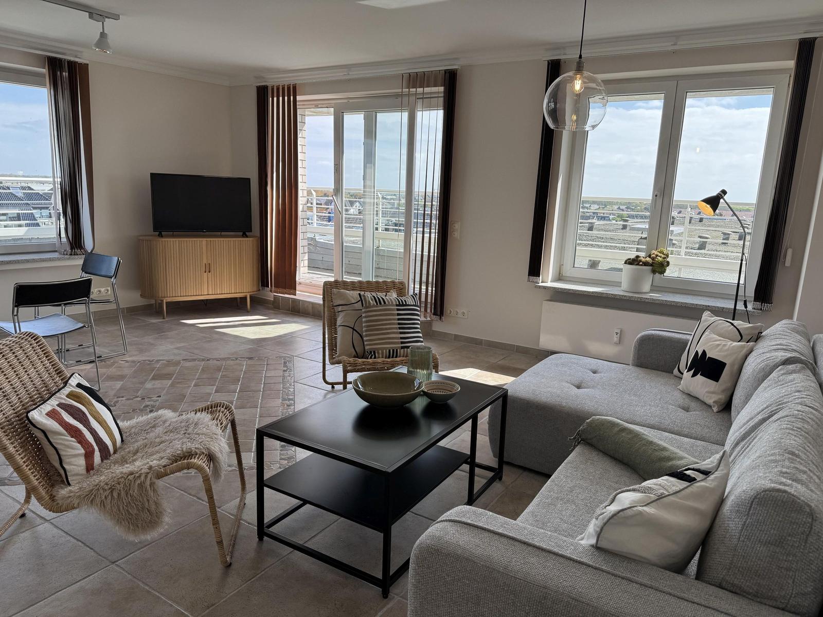 Living room with sofa, coffee table, and TV. Large windows with view of rooftops.