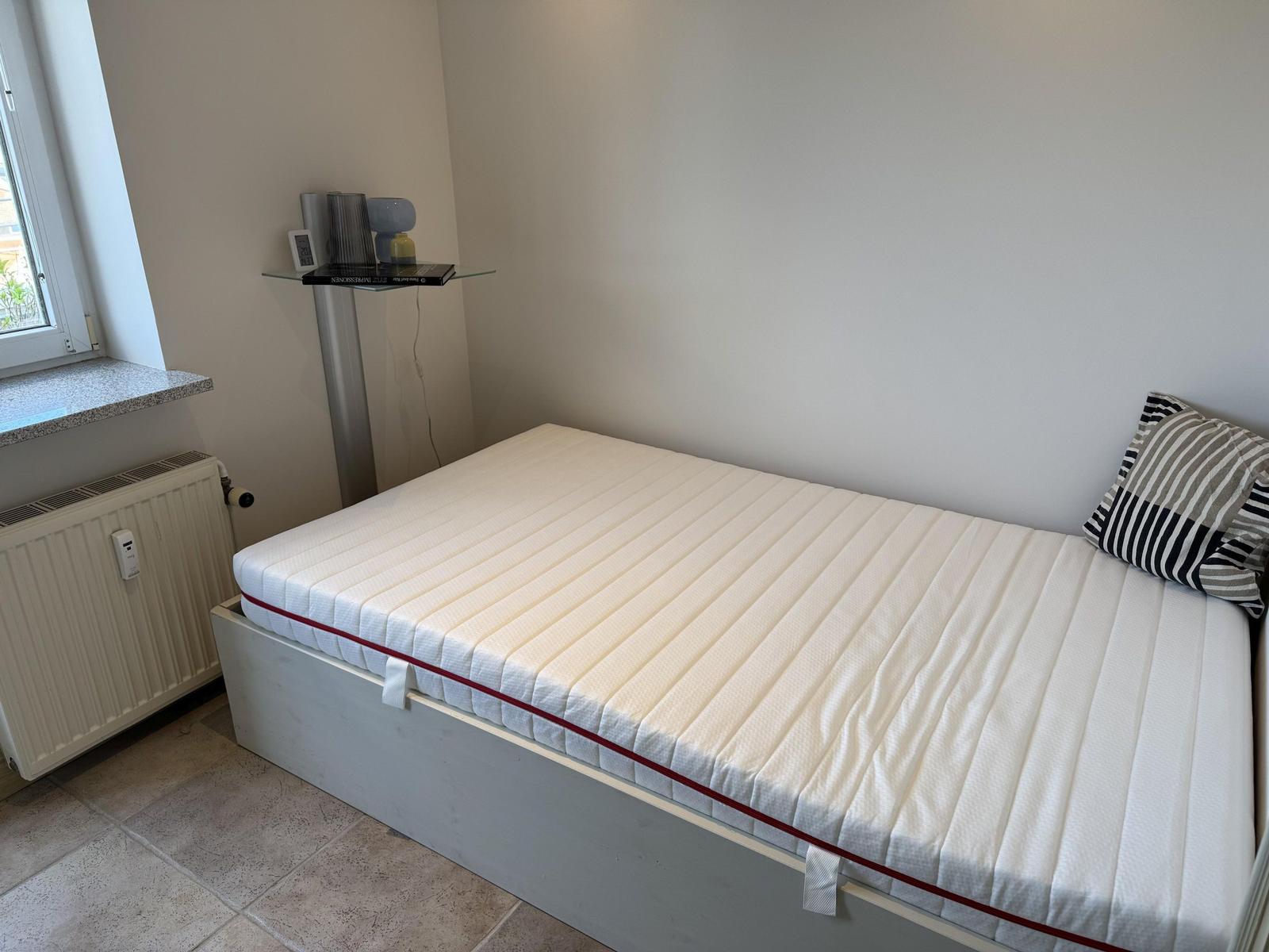 A bed with mattress and pillow in a room with window and radiator.