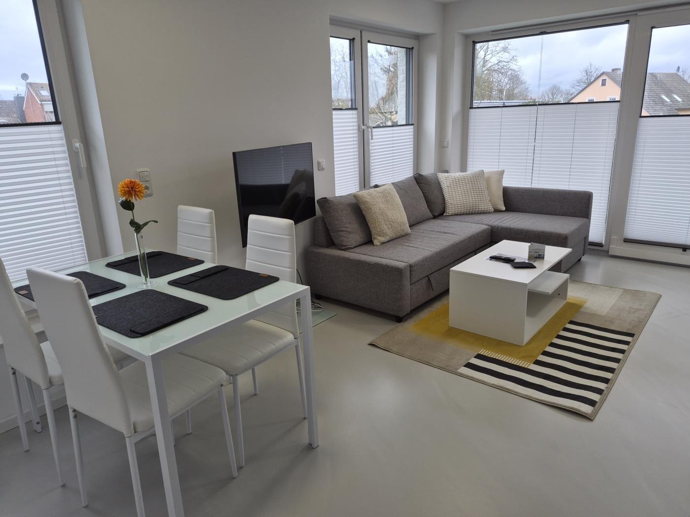 Wohn- und Essbereich mit Sofa, Tisch und Stühlen. Große Fenster mit Jalousien.