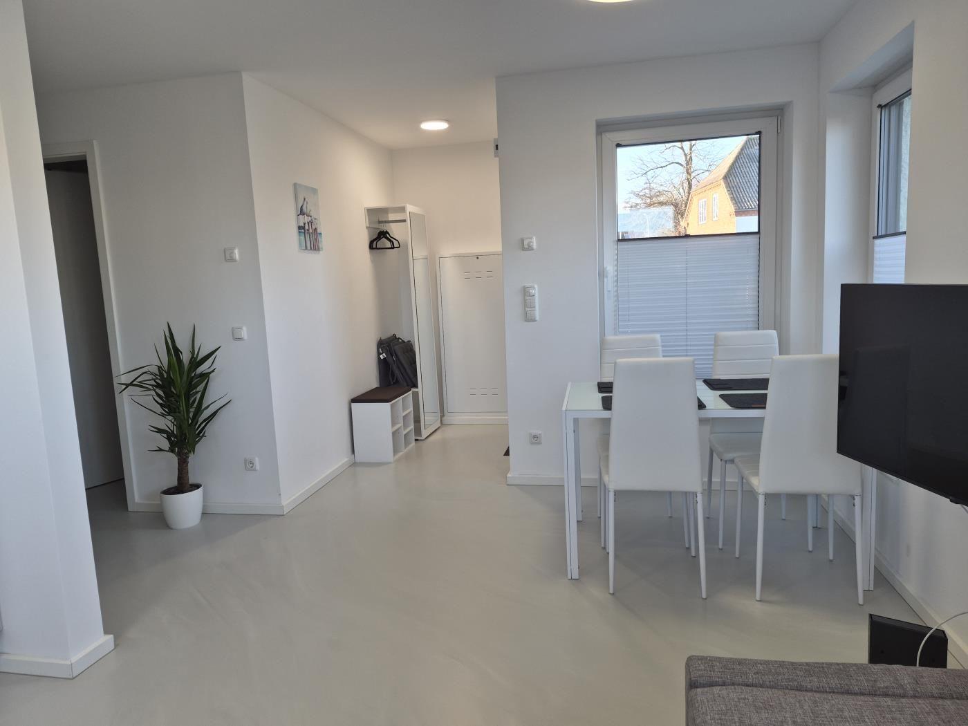 Wohn- und Essbereich mit weißem Tisch, Stühlen und Pflanze. Fenster mit Jalousie.