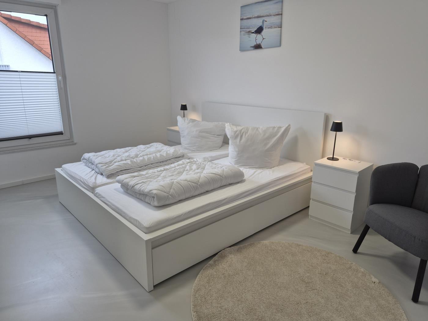 Schlafzimmer mit weißem Bett, Nachttischen und Stuhl. Fenster mit Jalousie und Wandbild.