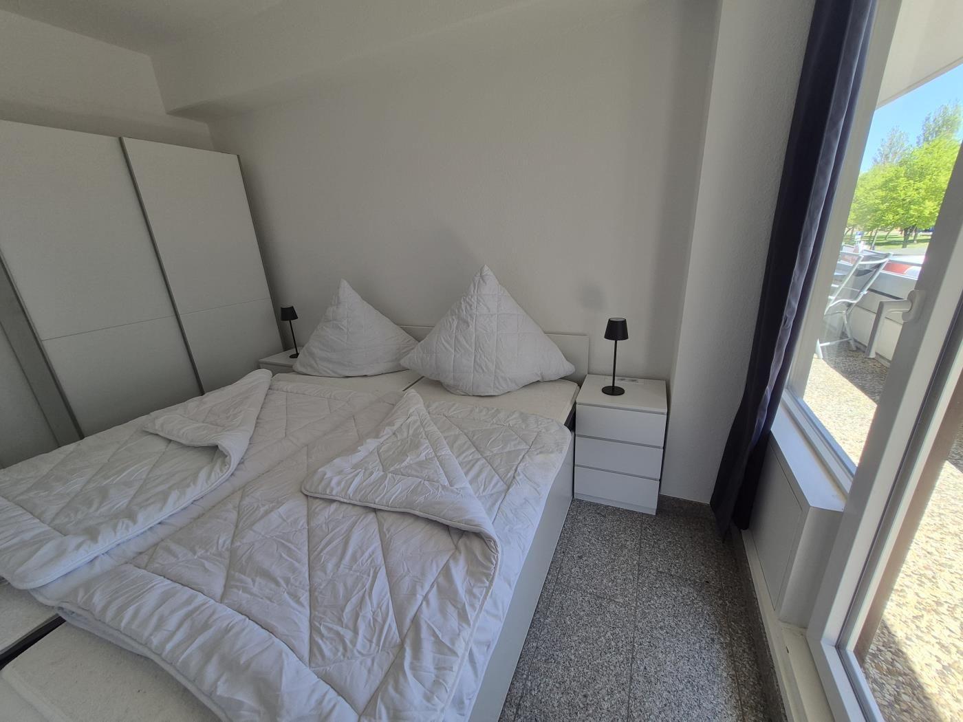 Schlafzimmer mit weißem Bett, Nachttischen und Fensterblick auf Garten.