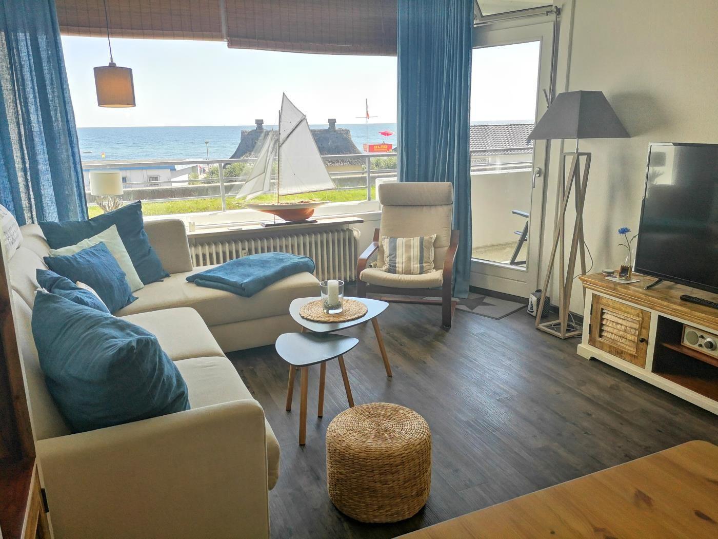 Wohnzimmer mit Sofa, Sessel und Fernseher. Blick auf Meer und Segelboot.