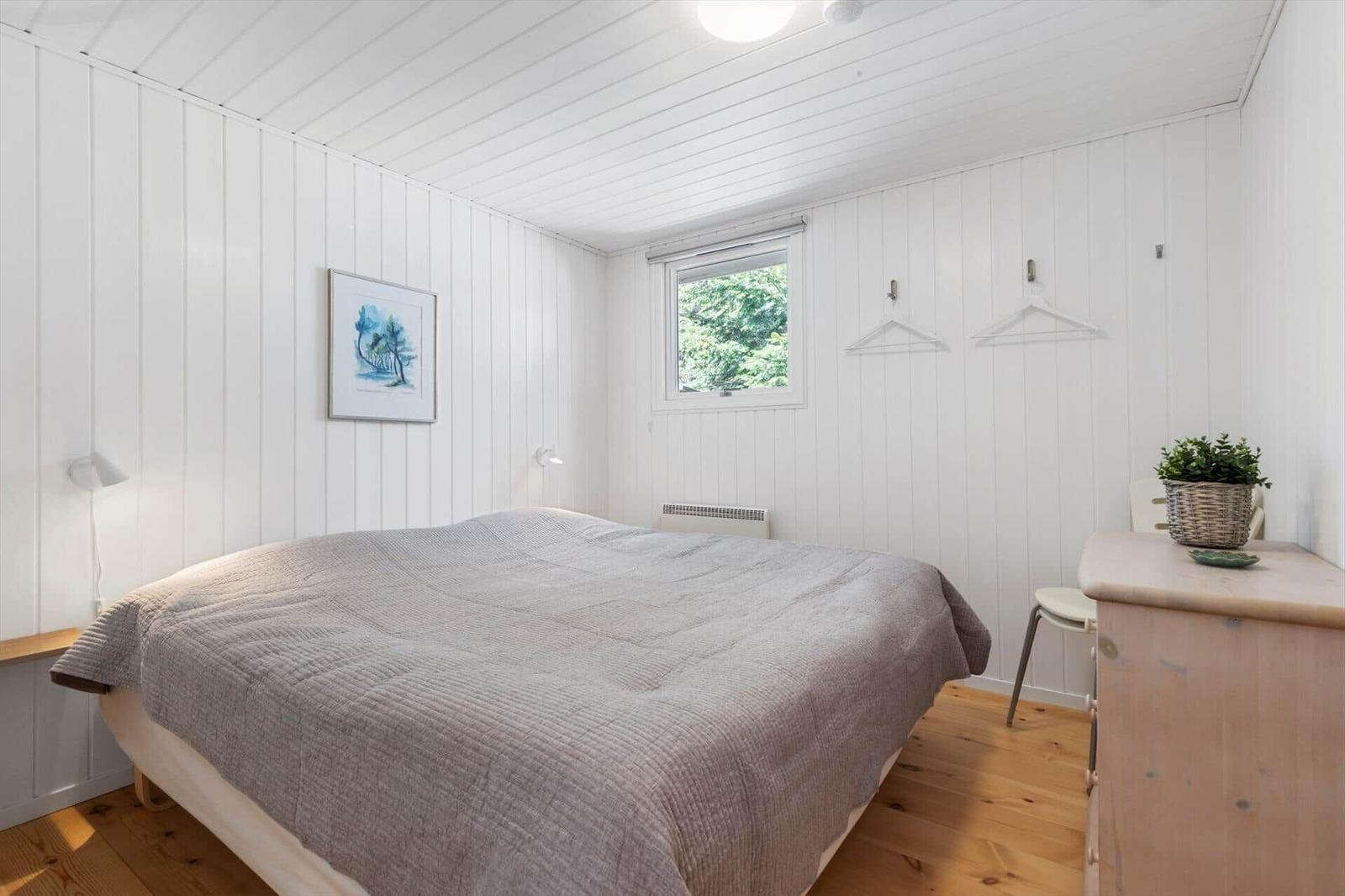Bedroom with bed, window, coat hooks, and dresser. Walls paneled white, wooden floor.