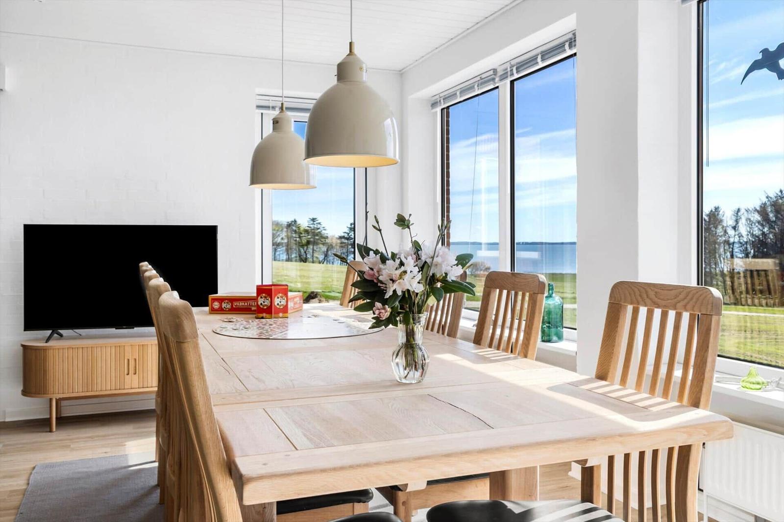 Dining room with wooden table, chairs, and sea view. Windows, TV, and flowers on table.