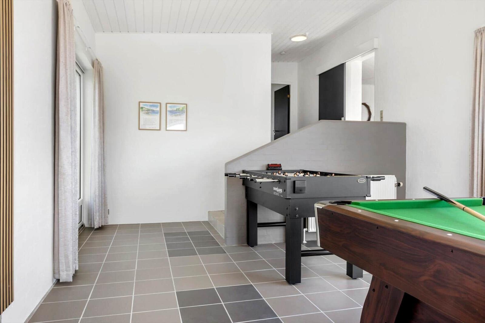 Room with pool table and foosball table, gray tiled floor and windows with curtains.