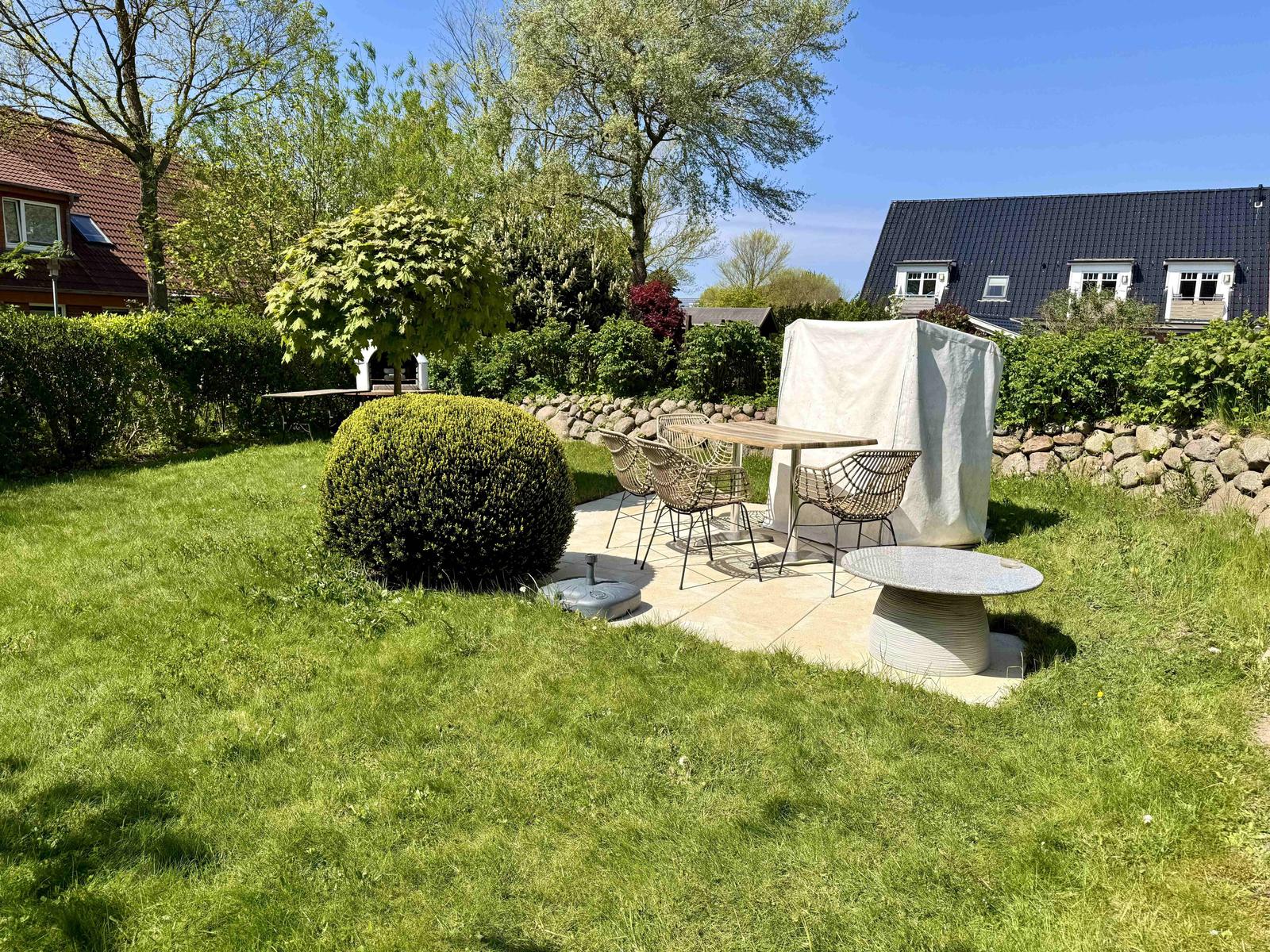 Garten mit Sitzbereich, Tisch, Stühlen und Beete. Hintergrund: Haus und Bäume.