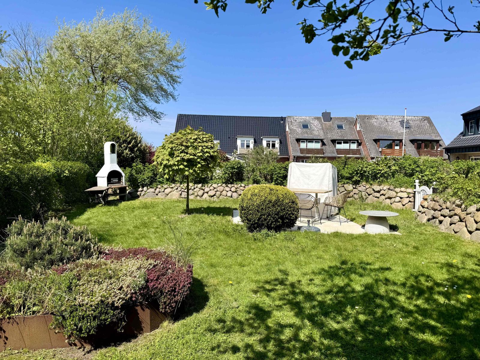 Garten mit Grill, Sitzgruppe und Blumenbeeten. Hintergrund: Wohnhäuser und Bäume.