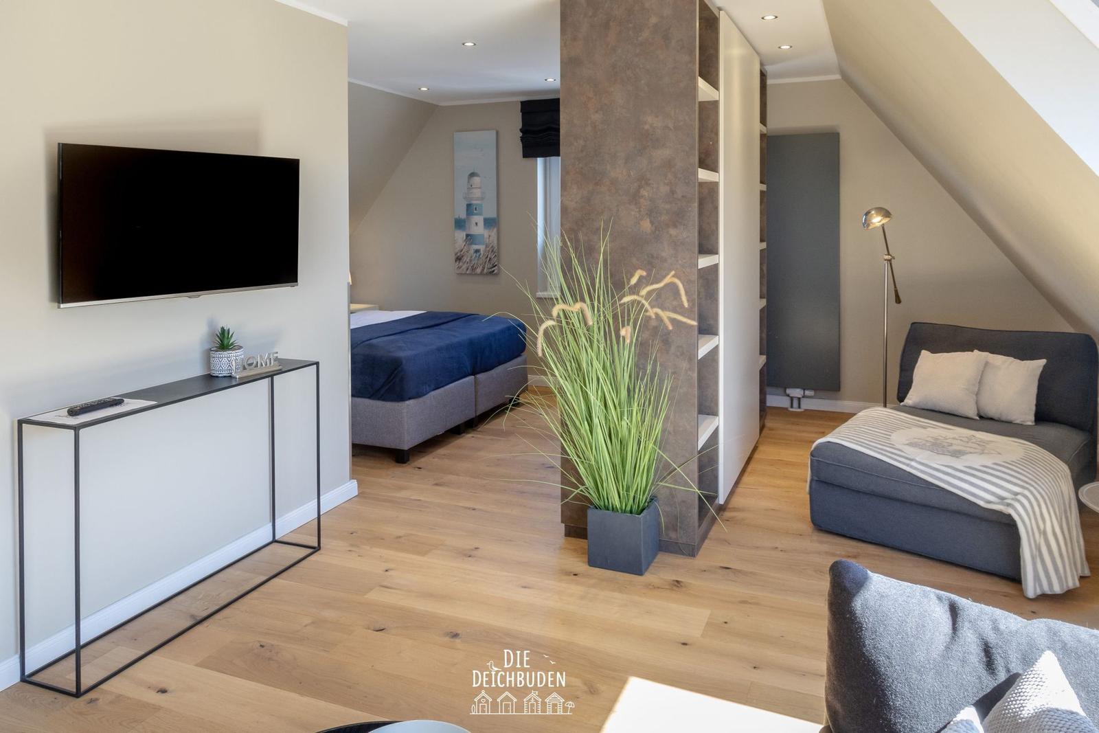Zimmer mit Schlafbereich, Fernseher und Pflanze. Holzboden und Dachschrägen.