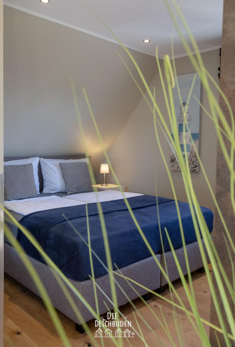 Schlafzimmer mit Doppelbett, Wandbild Leuchtturm und Stehgras im Vordergrund.