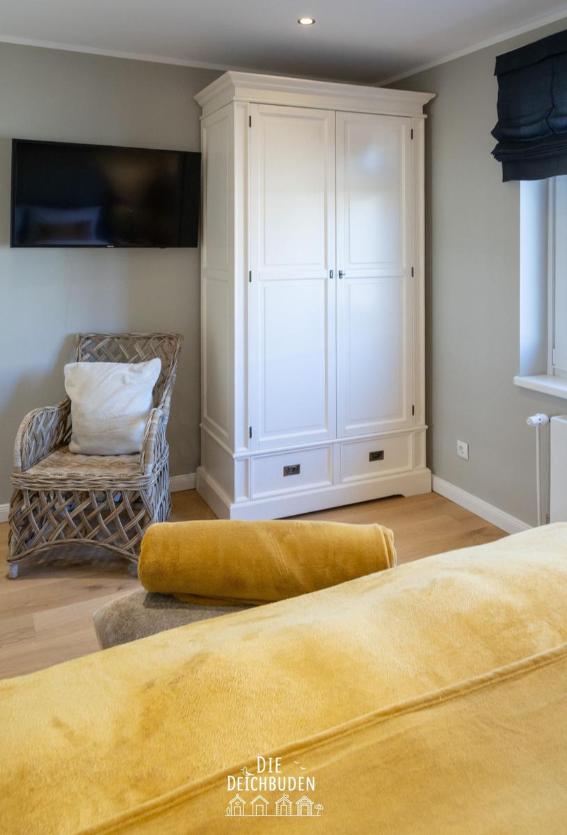 Schlafzimmer mit weißem Schrank, TV und geflochtenem Sessel.