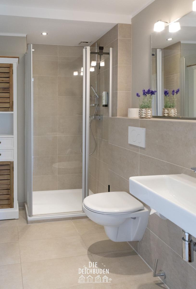 Duschbereich mit Duschkabine, Toilette und Waschbecken. Holzregal und Lavendelblumen.