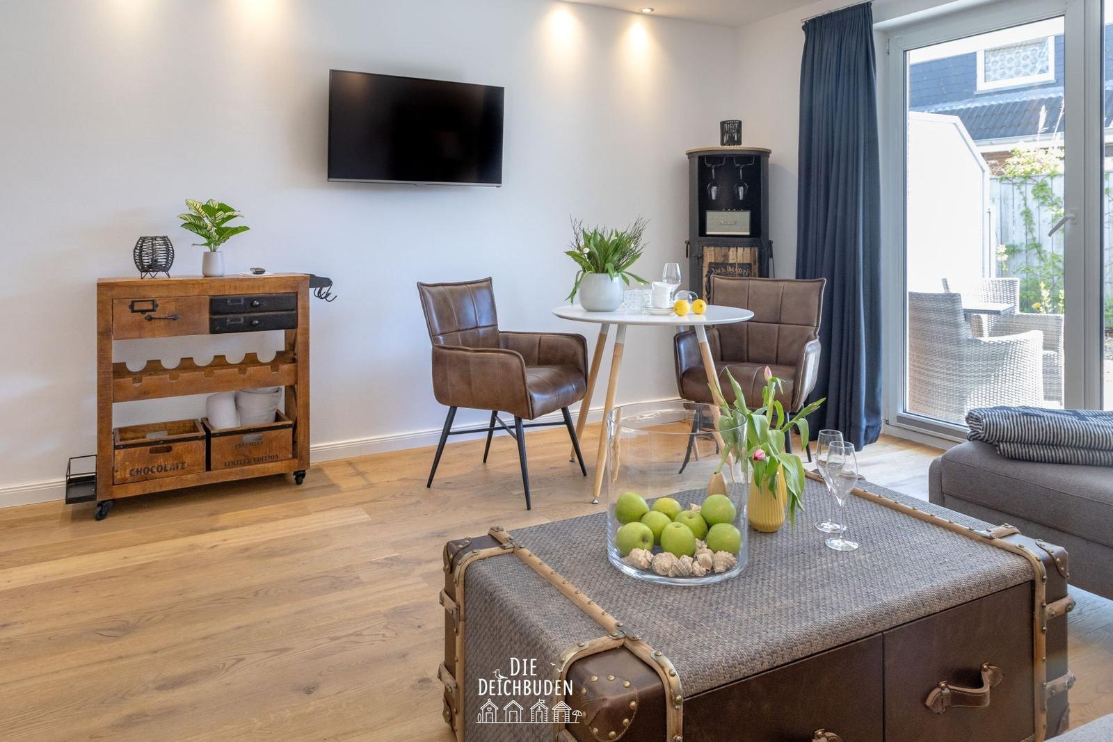Wohnzimmer mit Holzboden, Fernseher, Tisch und Stühlen, Koffer-Tisch und Blick auf Terrasse.