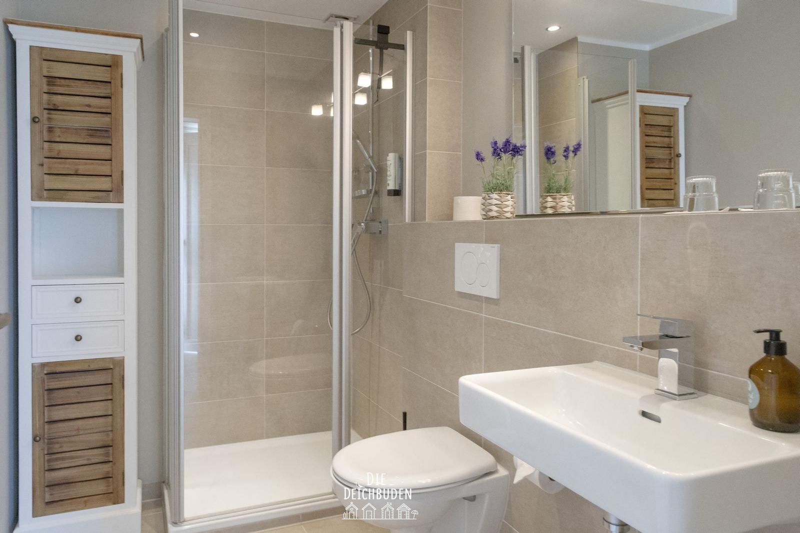 Badezimmer mit Dusche, Waschbecken und Toilette. Holzschrank und Lavendelblumen.