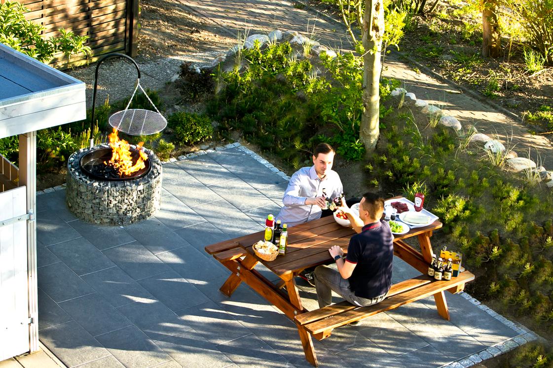 Zwei Personen sitzen an einem Holztisch auf einer Terrasse mit Feuerkorb und Grillzange.