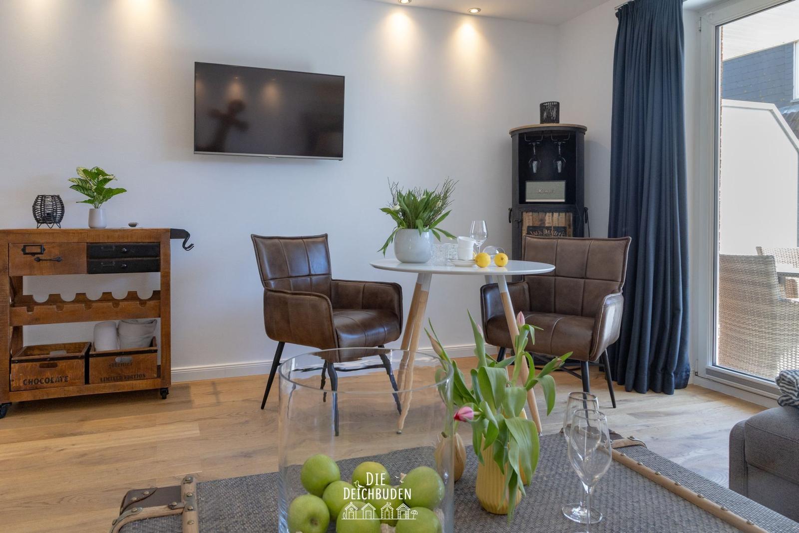Wohnzimmer mit TV, Esstisch und Sitzgelegenheiten. Holzboden und große Fenster.