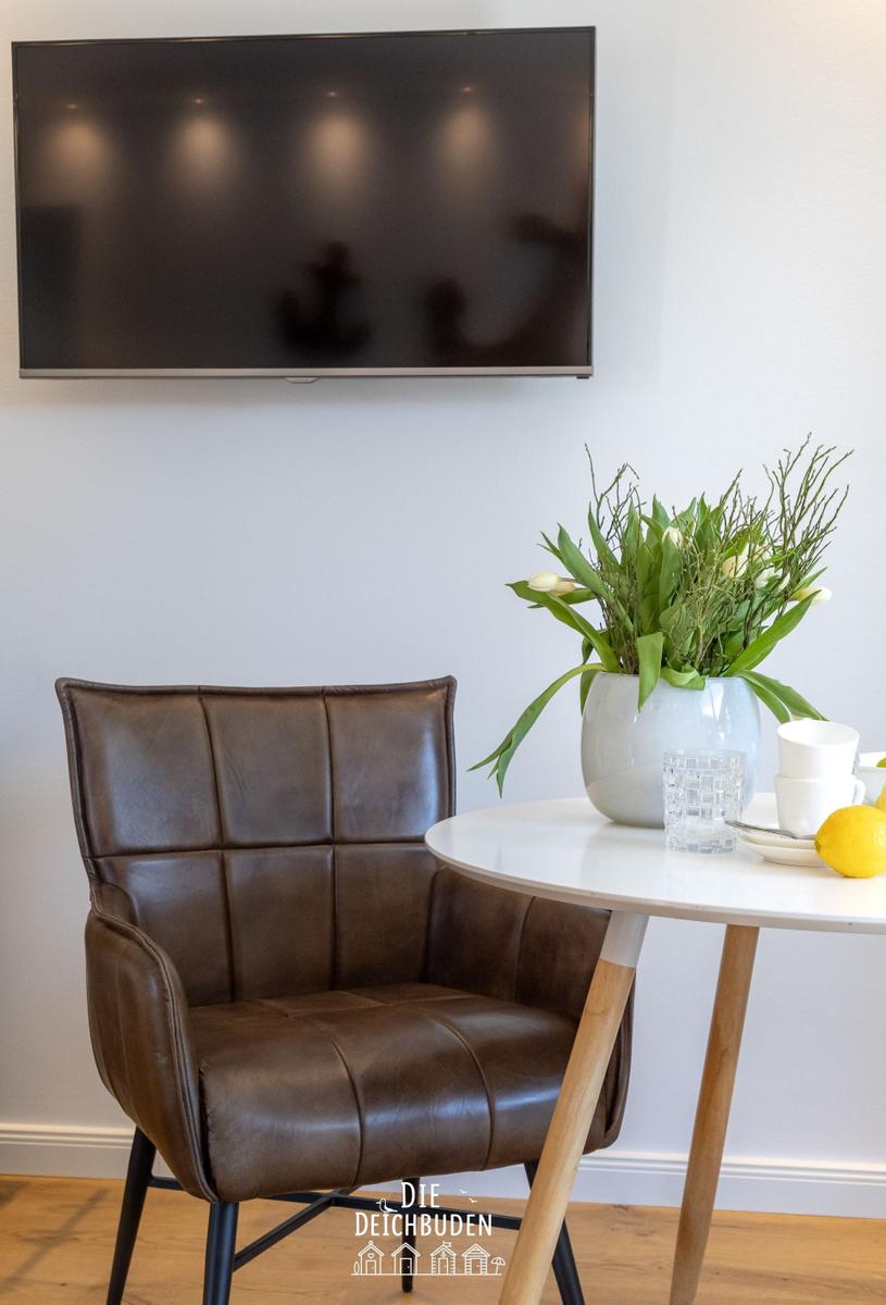 Ein braunes Ledersofa steht vor einem weißen Tisch mit Blumen und Gläsern. Über dem Sofa hängt ein Flachbildschirm.