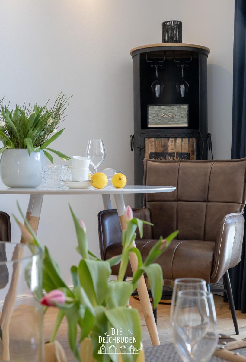 Esstisch mit Stühlen, Blumen und Weinflaschenhalter im Hintergrund.