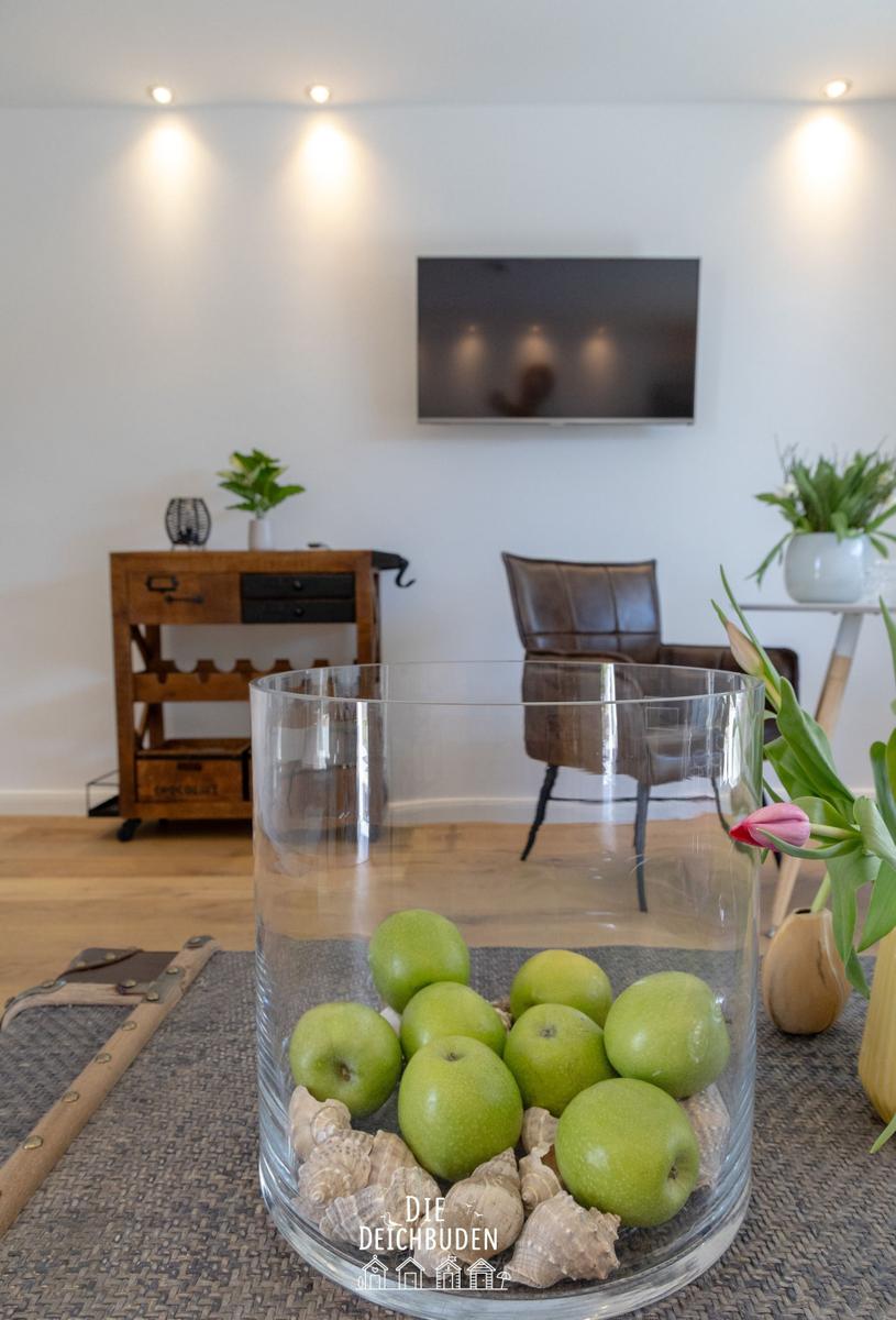 Ein Glas mit grünen Äpfeln und Muscheln steht auf einem Tisch. Im Hintergrund ein Fernseher und ein Holzregal.