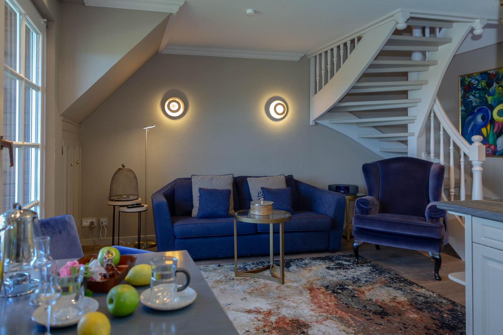 Ein Wohnzimmer mit blauem Sofa, Sessel und Treppe. Auf dem Tisch stehen Obst und Gläser.